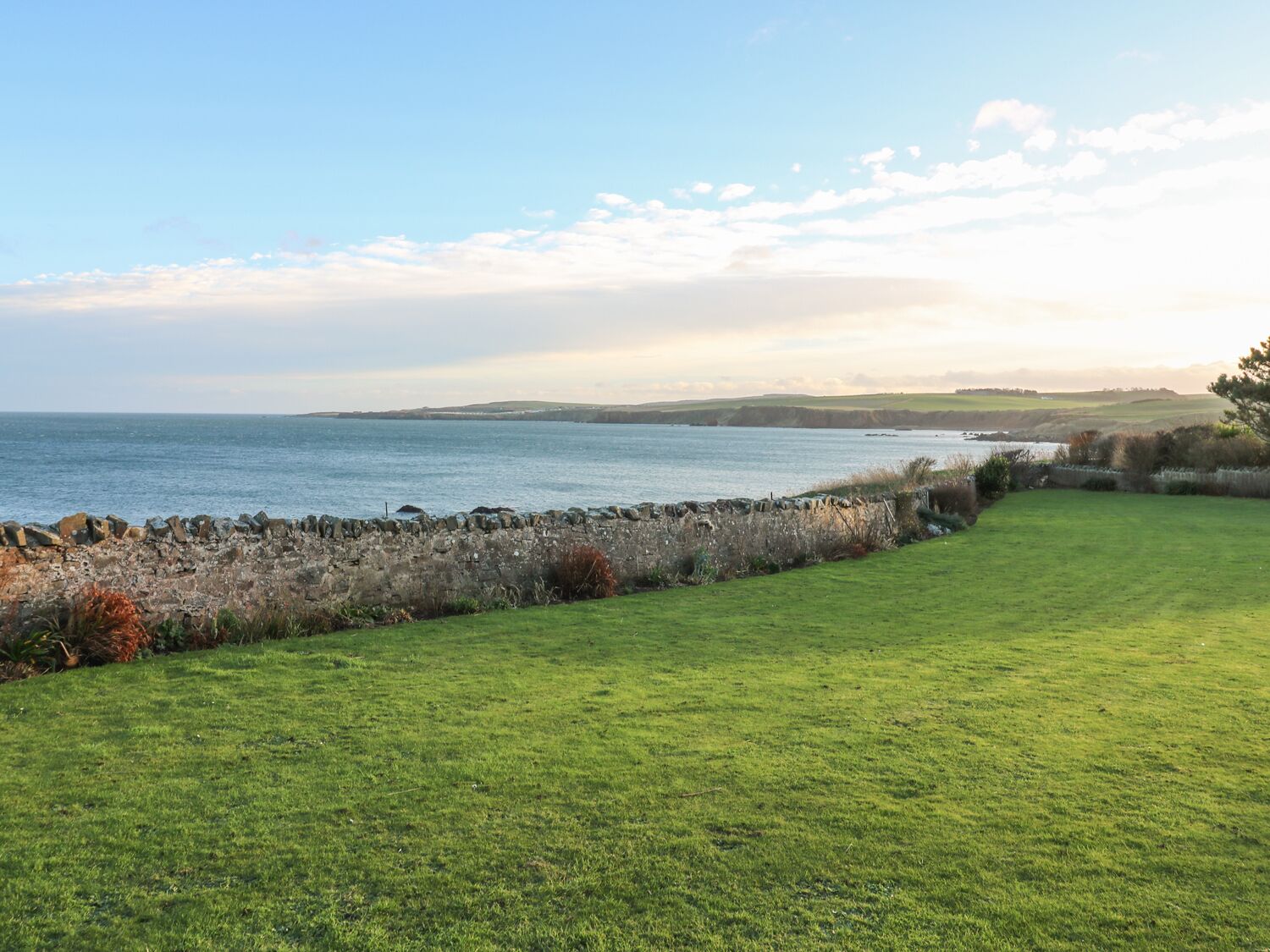 A coastal landscape with a grassy area, stone wall, and a view of the sea and distant hills.