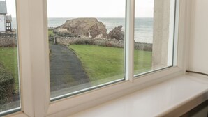 Blick durch ein Fenster auf die Küste, mit einem Grasweg, einem Felsen und dem Meer.