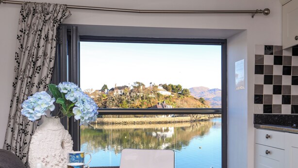 A dining area with a table set for two, a vase of flowers, and a view of a lake and houses in the background. - St Agnes (Porthmadog)