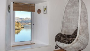 A room with a large window offering a view of a lake and mountains, a hanging chair, and a small wooden table. - St Agnes (Porthmadog)