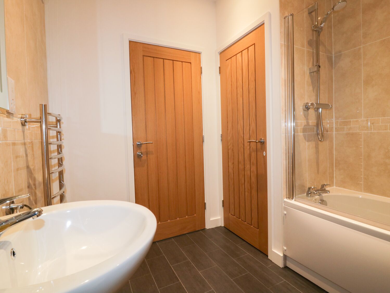 Une salle de bain avec un grand lavabo blanc, une baignoire et une douche. On y trouve deux portes en bois et un porte-serviettes.