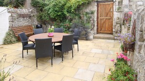 A stone-walled patio with a round wooden table and chairs, surrounded by potted plants and flowers.