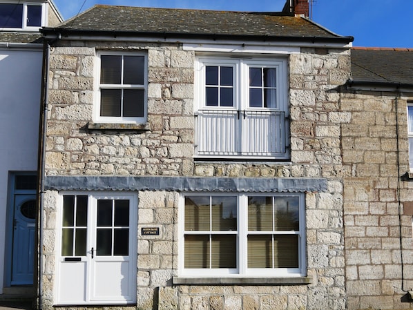 A stone building with two windows, a door, and a sign that reads "Alpine Rose Cottage."