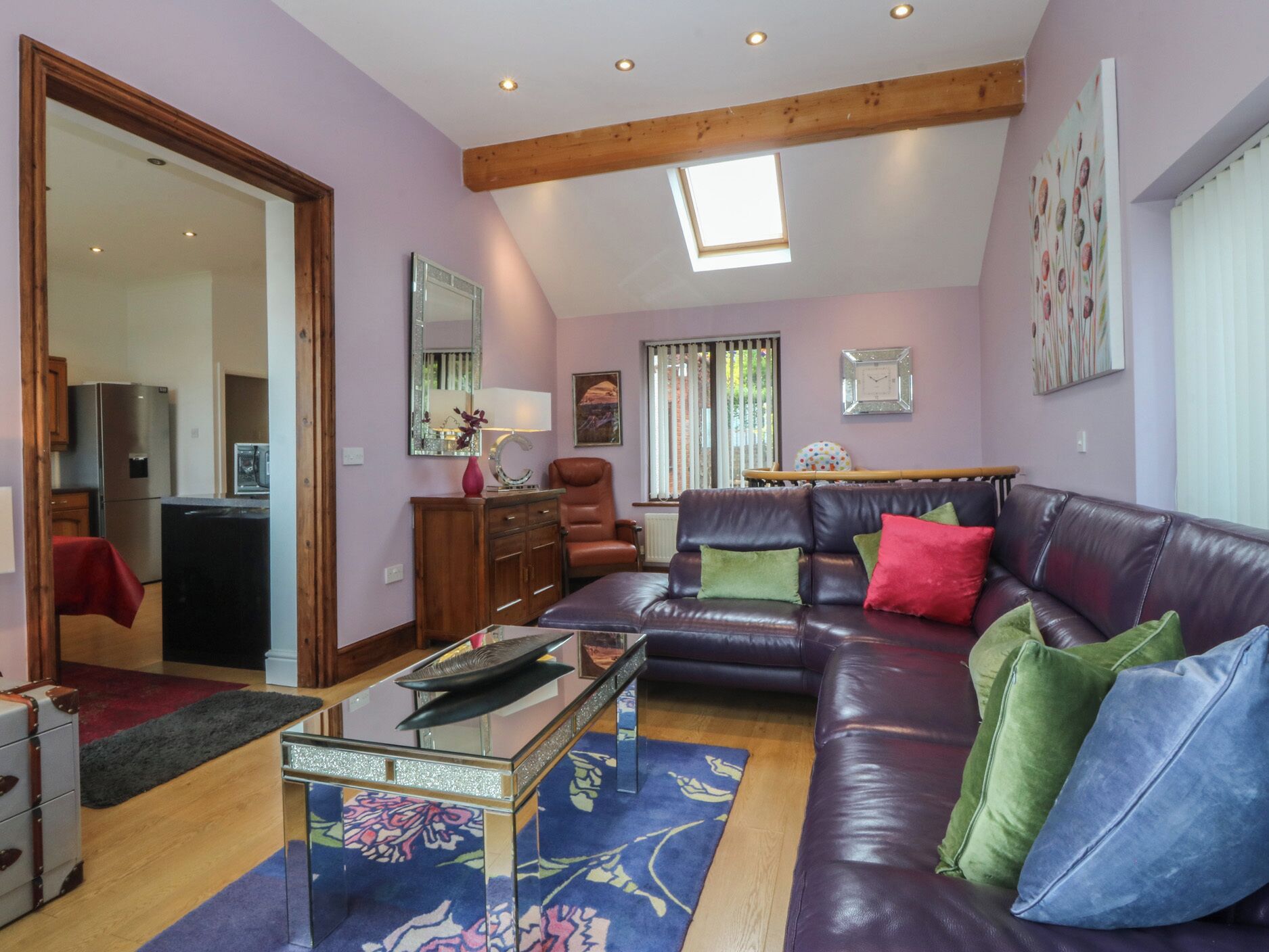 Un salon avec un canapé en cuir, une table basse en verre, un meuble en bois, un fauteuil rouge et un coin cuisine visible à travers une porte ouverte.