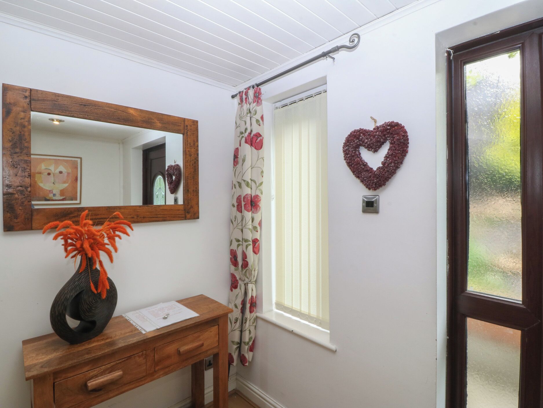 A room with a wooden desk, a mirror, a heart-shaped decoration, a window, and a door.