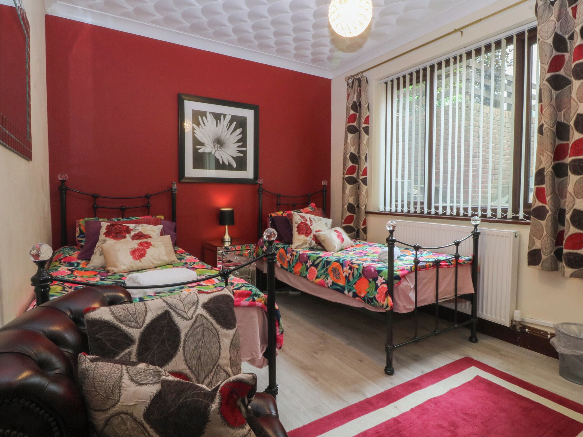 A bedroom with two beds, a red wall, a framed flower, and a window with curtains.