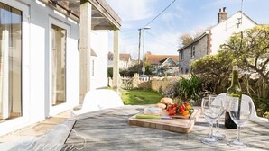 A wooden table with a cheese board, a bottle of wine, and glasses, set on a patio with a view of a white house and a garden. - Blueberry Cottage (Weymouth)