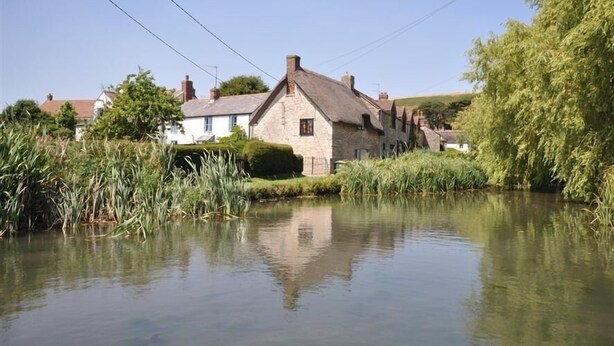 A stone house with a thatched roof by a calm river, surrounded by greenery and other buildings. - Blueberry Cottage (Weymouth)