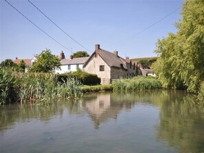 Une maison en pierre avec un toit de chaume, située au bord d’une rivière paisible, entourée de verdure et d’autres bâtiments.