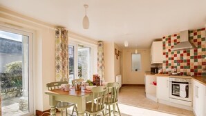 A dining area with a table and chairs, a kitchen with a tiled backsplash, and a view of the outside. - Blueberry Cottage (Weymouth)