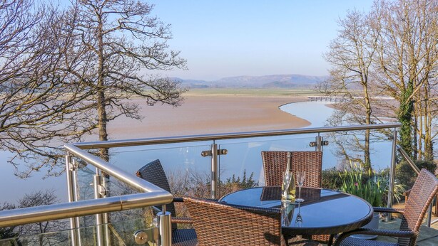A glass-enclosed outdoor seating area with wicker furniture overlooking a river. - Shady Bowers (Carnforth)