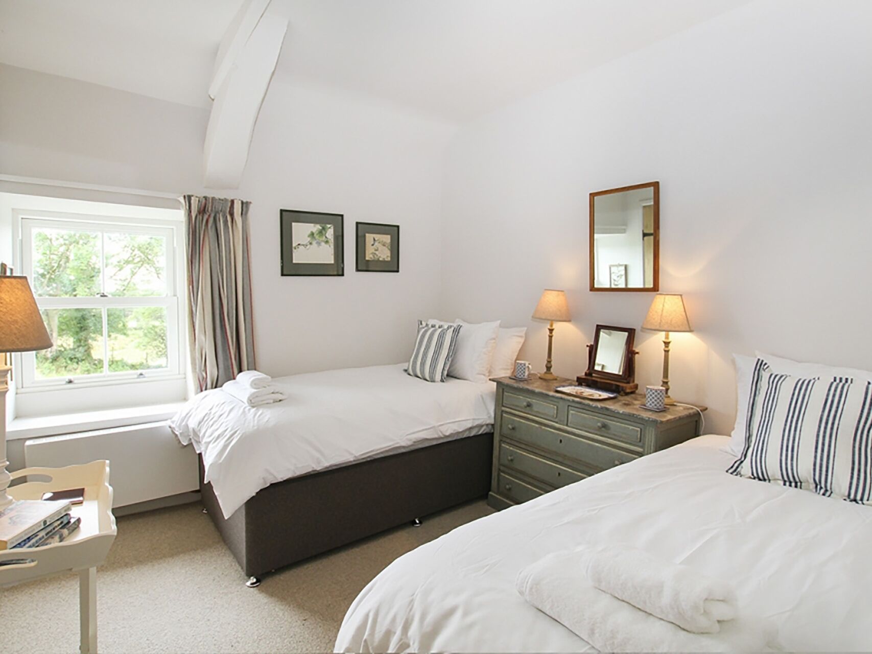 A bedroom with two beds, a dresser, a window with curtains, and framed artwork on the wall.