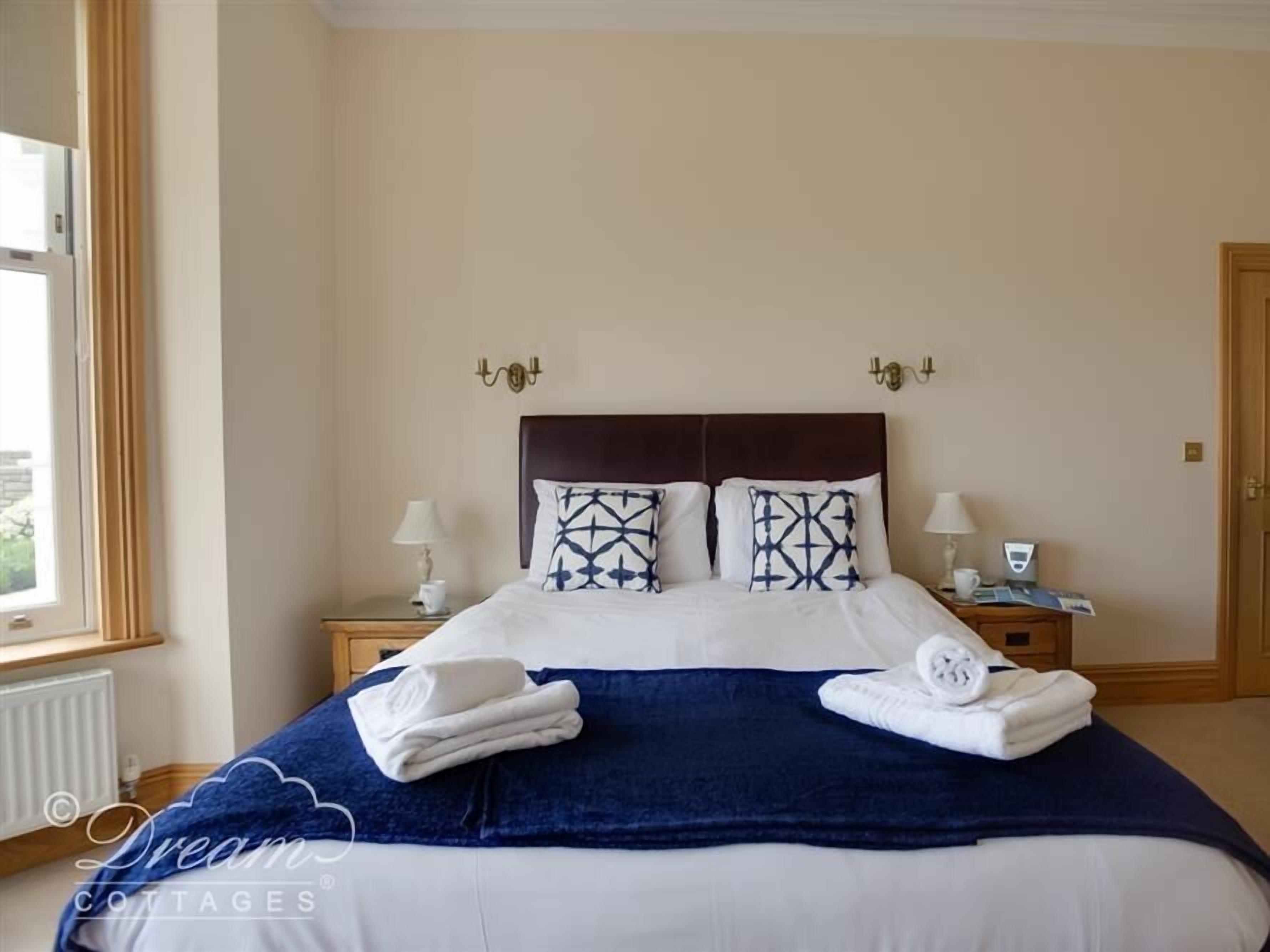 Una cama bien hecha con una manta azul oscuro y almohadas blancas, dos lámparas de noche, una ventana con cortinas y una puerta.