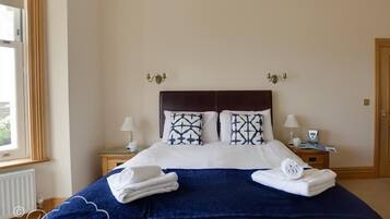Una cama bien hecha con una manta azul oscuro y almohadas blancas, dos lámparas de noche, una ventana con cortinas y una puerta.