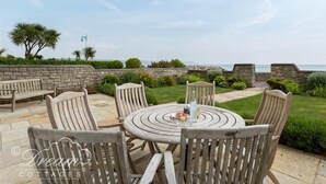 Un espace extérieur aménagé avec des meubles en bois, comprenant une table ronde et des chaises, offrant une vue sur un jardin paysager et l’océan.