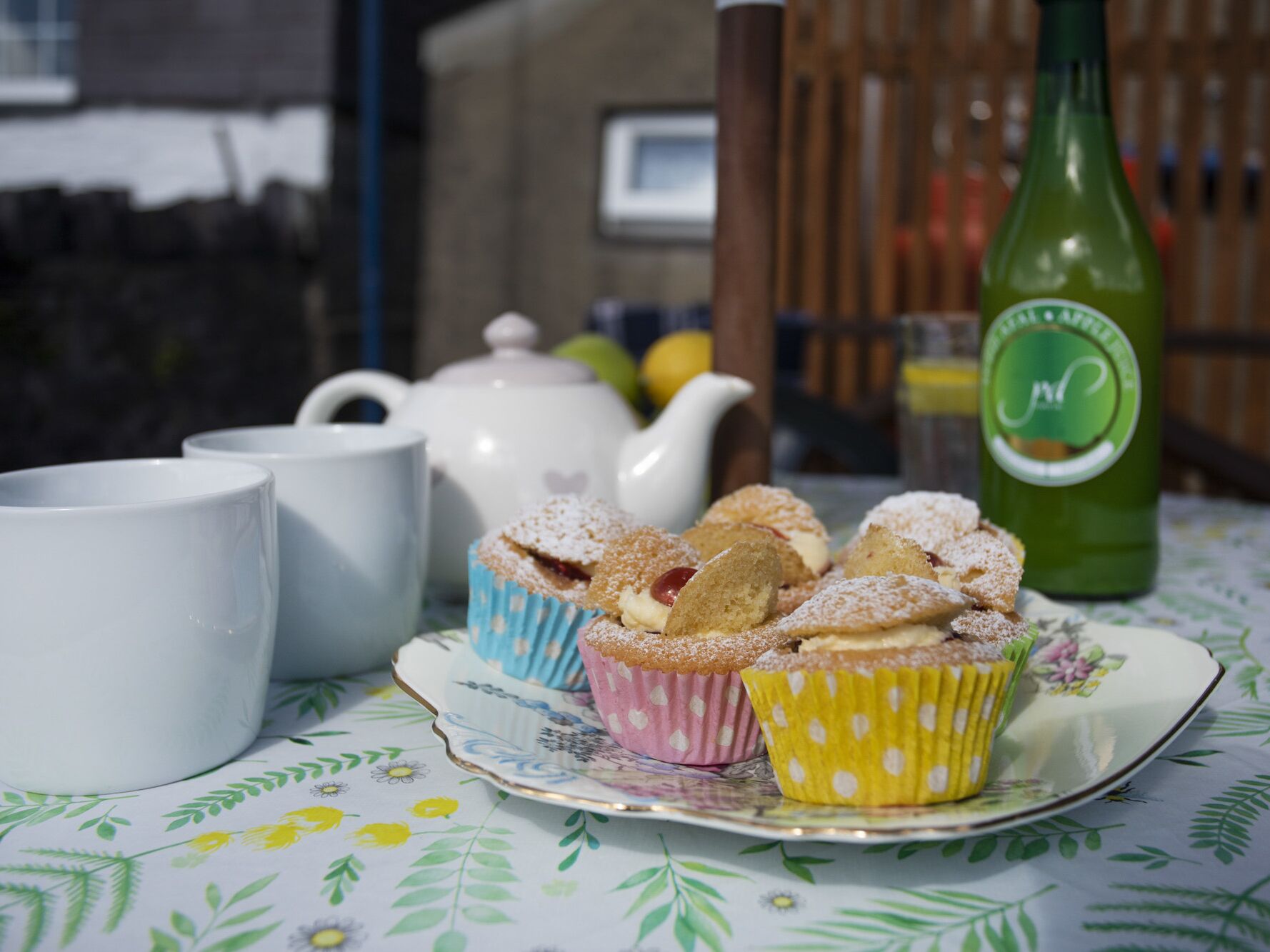 Une table recouverte d’une nappe à motifs floraux, une théière, deux tasses blanches, une assiette avec des petits gâteaux et une bouteille verte.