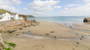 A coastal scene with a sandy beach, a white building with a sloped roof, and a rocky cliff. - Brambles (St Austell)