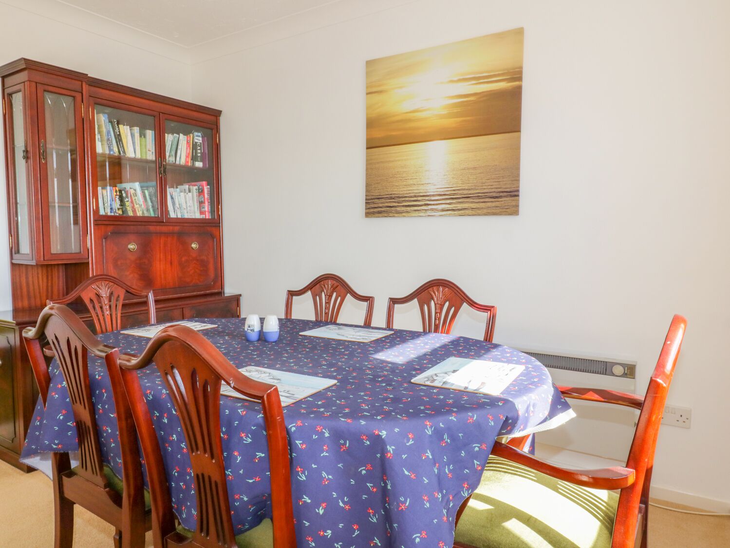 A dining area with a round table, four chairs, a wooden cabinet with glass doors, and a sunset painting on the wall.