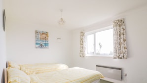 A bedroom with a bed, a radiator, a window with curtains, and a wall-mounted picture. - Brambles (St Austell)