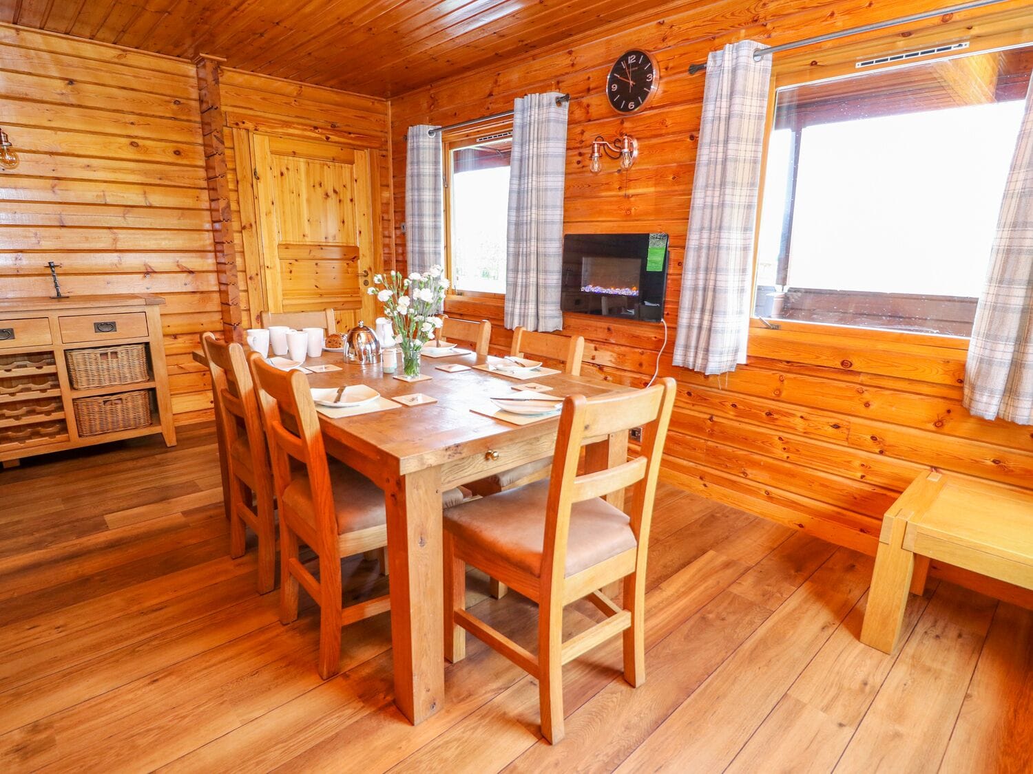 A wooden cabin with a dining area featuring a wooden table and chairs, a television, and plaid curtains.