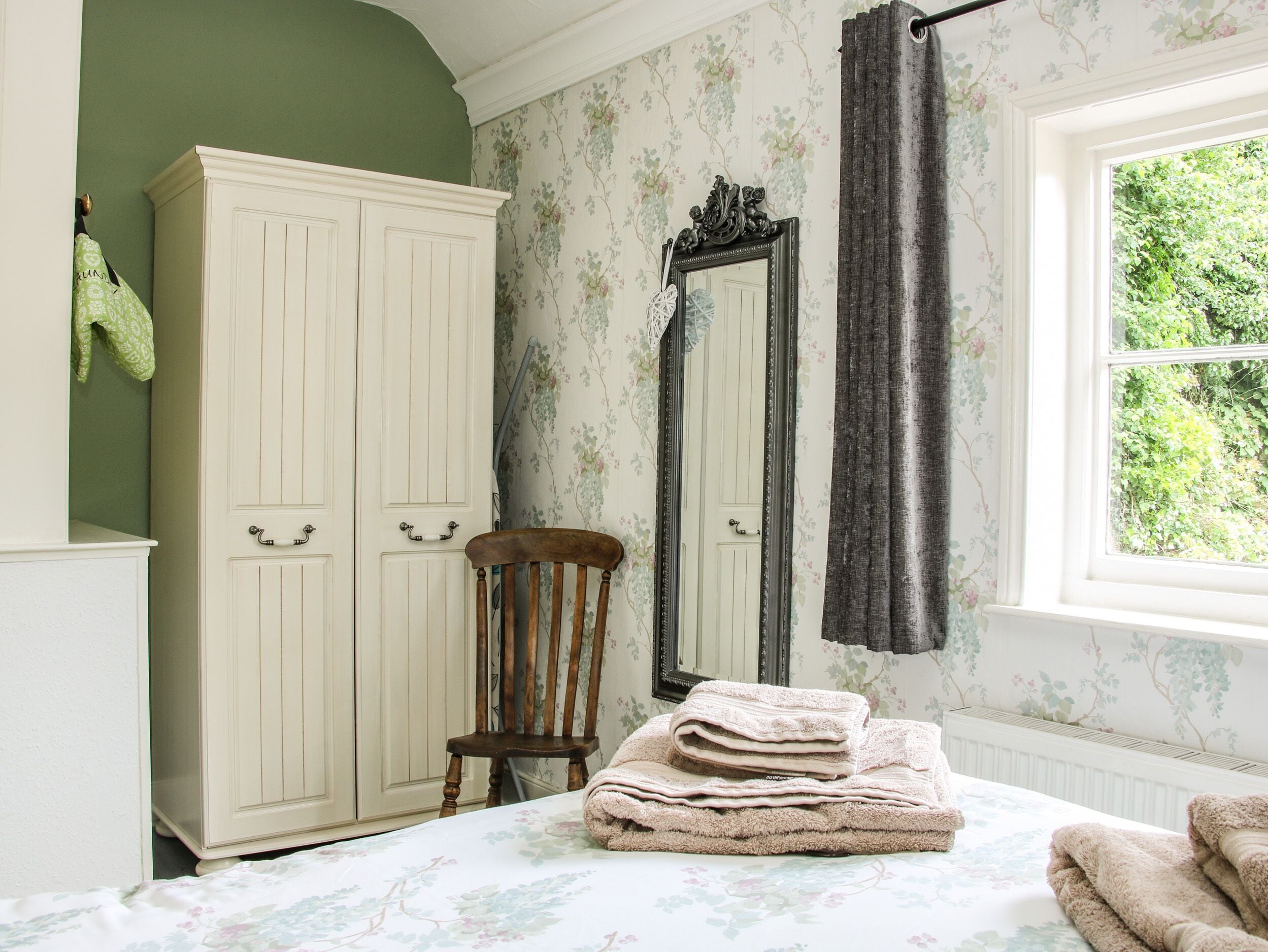 A bedroom with a bed, a wardrobe, a chair, a mirror, and a window with a view of greenery.