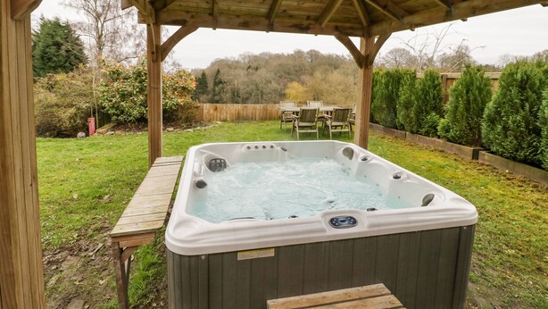 A hot tub under a wooden gazebo in a backyard with a wooden table and chairs, surrounded by greenery and a wooden fence. - Shatterford Lakes (Bewdley)