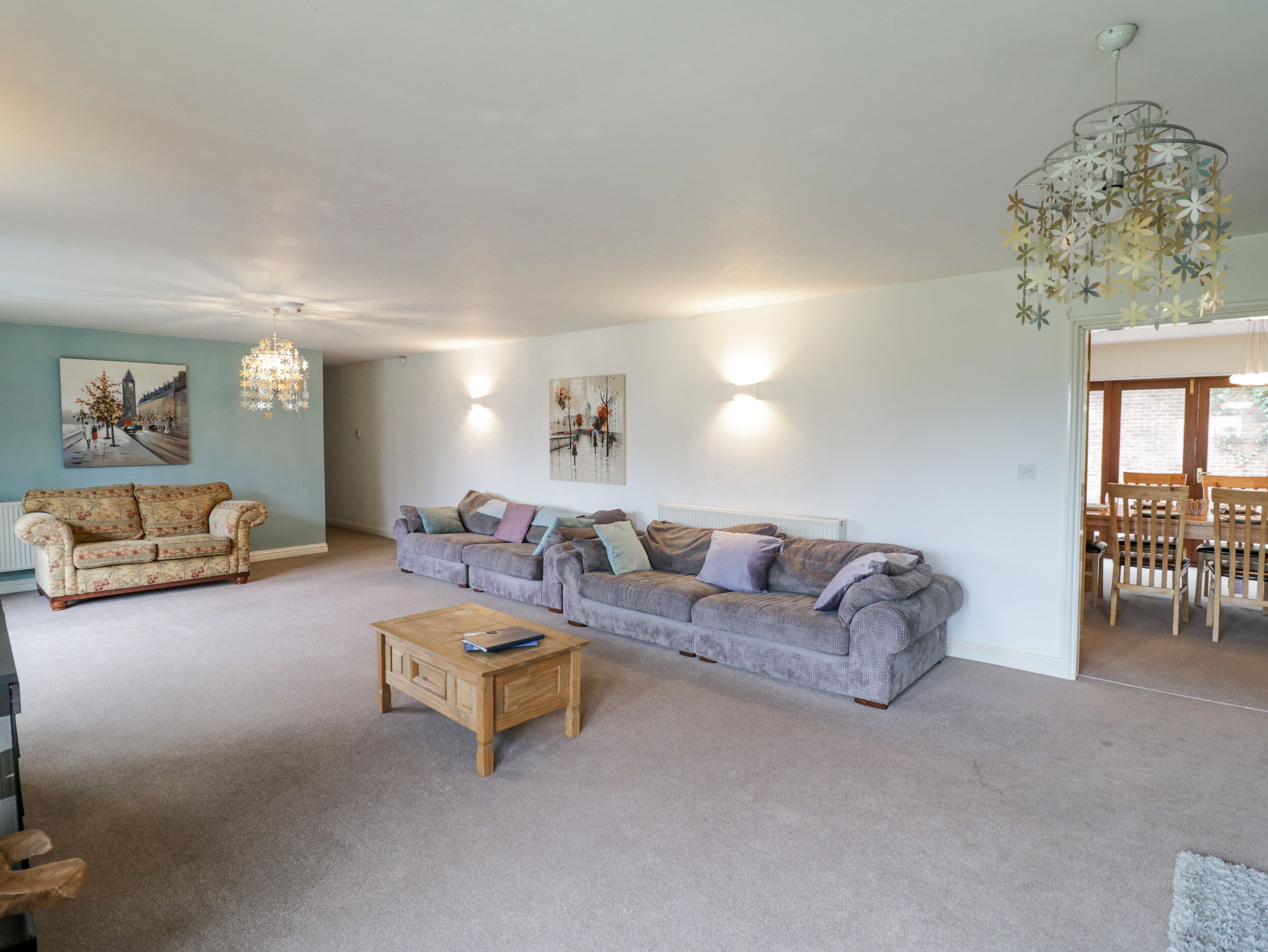 A spacious living room with a large grey sofa set, a wooden coffee table, and a chandelier. There are two wall-mounted lights and a painting on the wall.