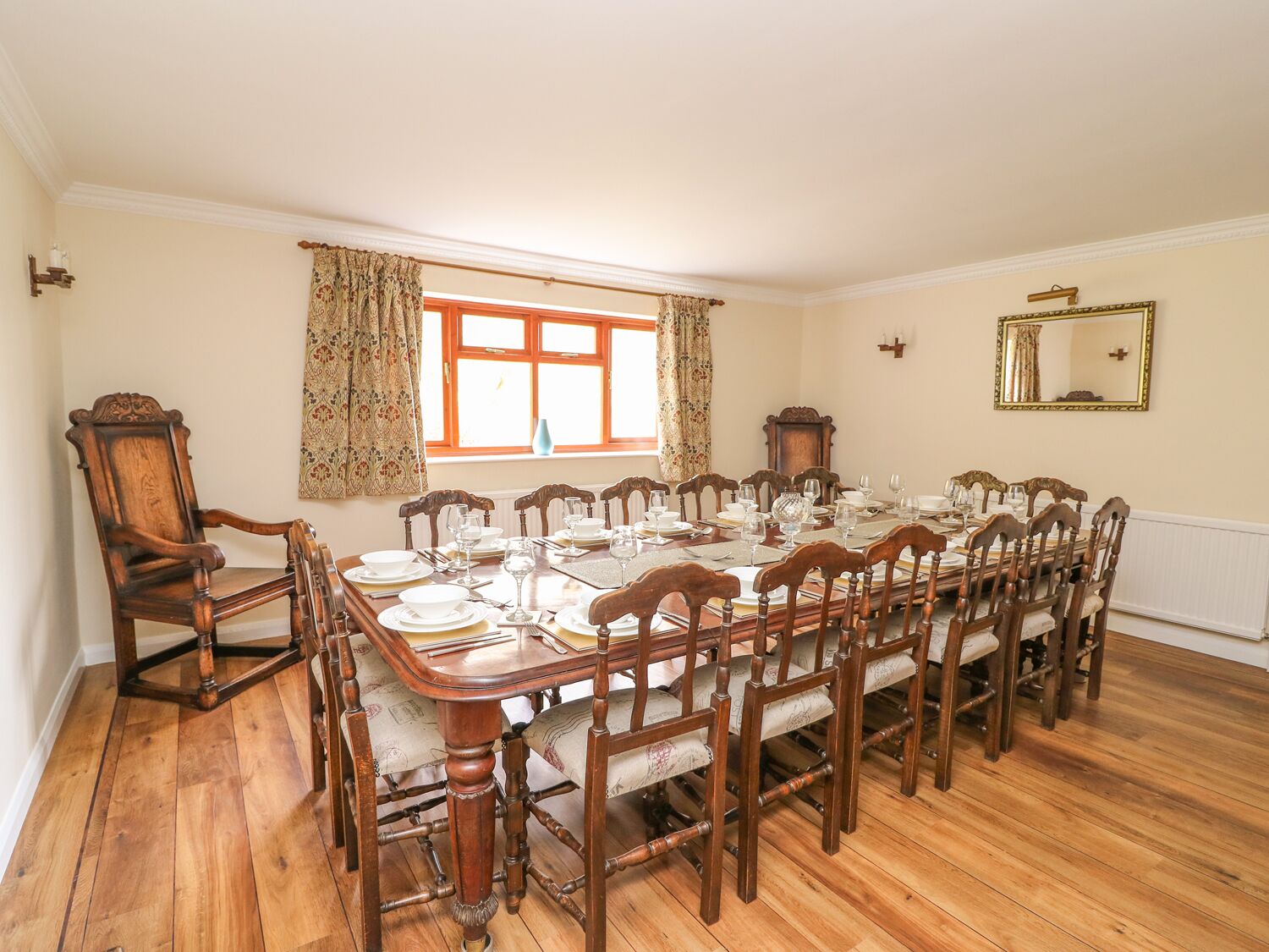 A dining room with a long wooden table set for a meal, flanked by wooden chairs, a window with curtains, and a mirror on the wall.