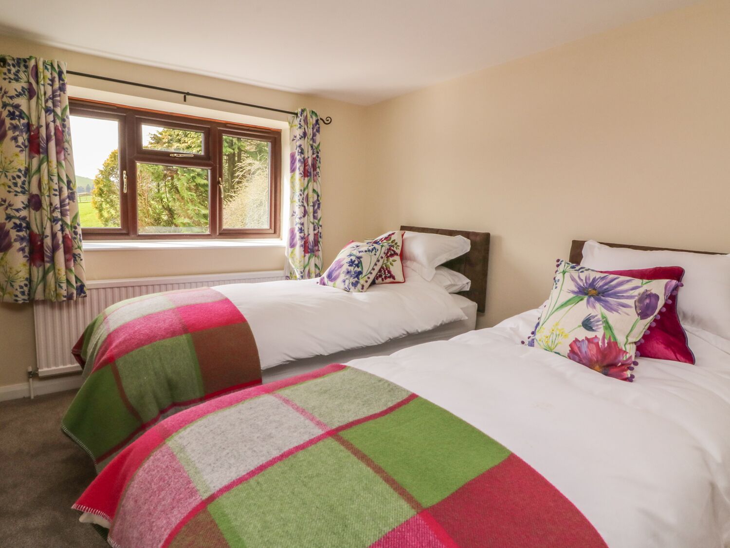 A bedroom with two beds, floral curtains, and a window with a view of greenery.