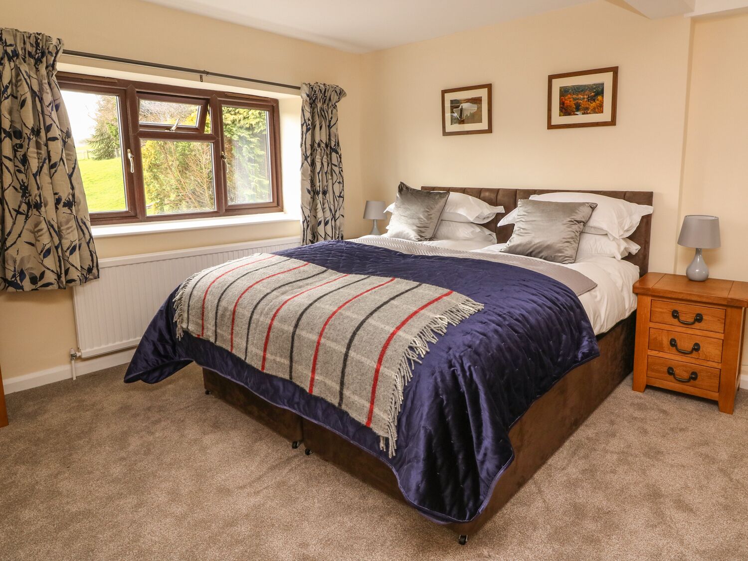 A bedroom with a bed, a nightstand, a window with curtains, and two framed pictures on the wall.