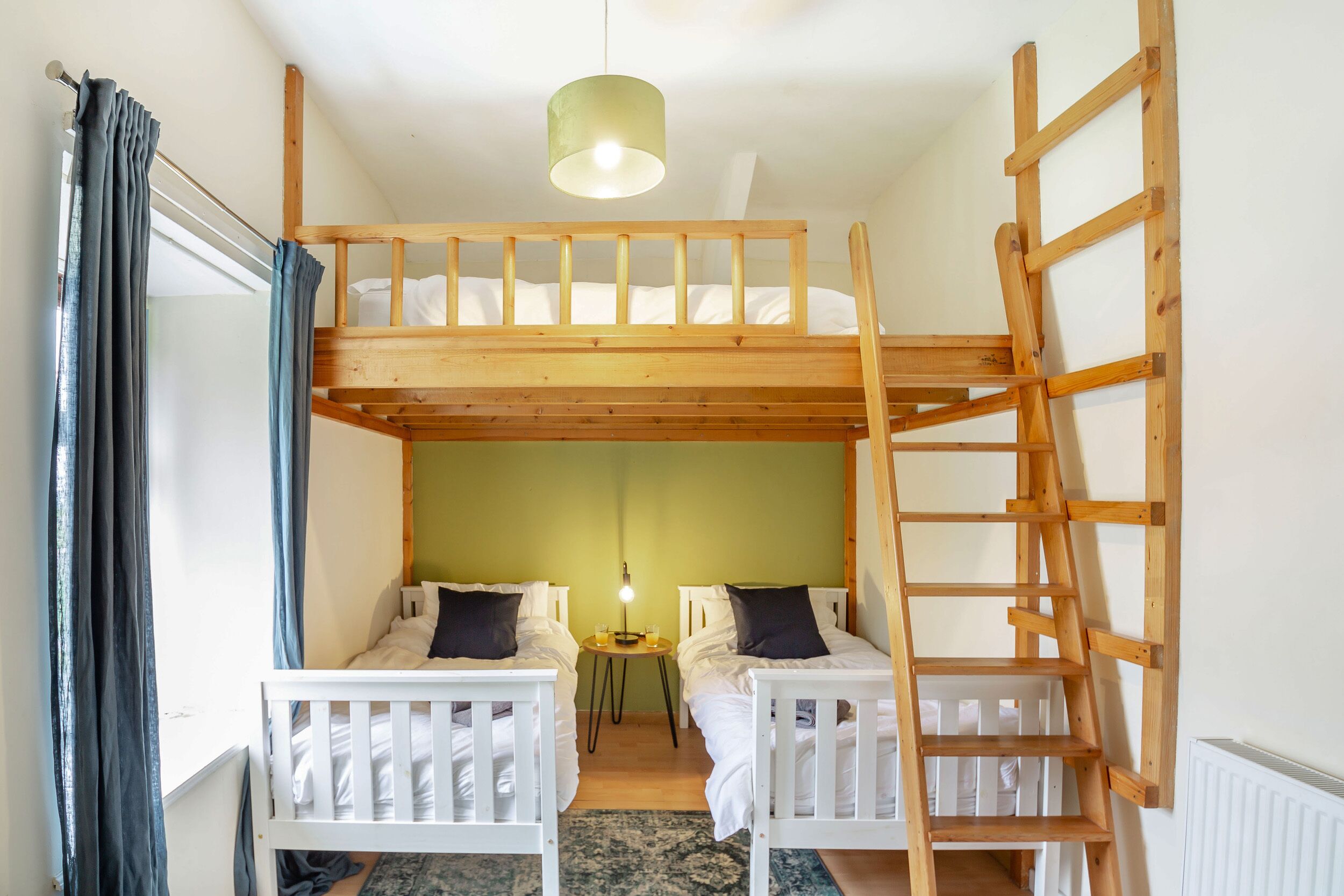 A dormitory room with bunk beds, a ladder, a small table with a lamp, and a window with curtains.