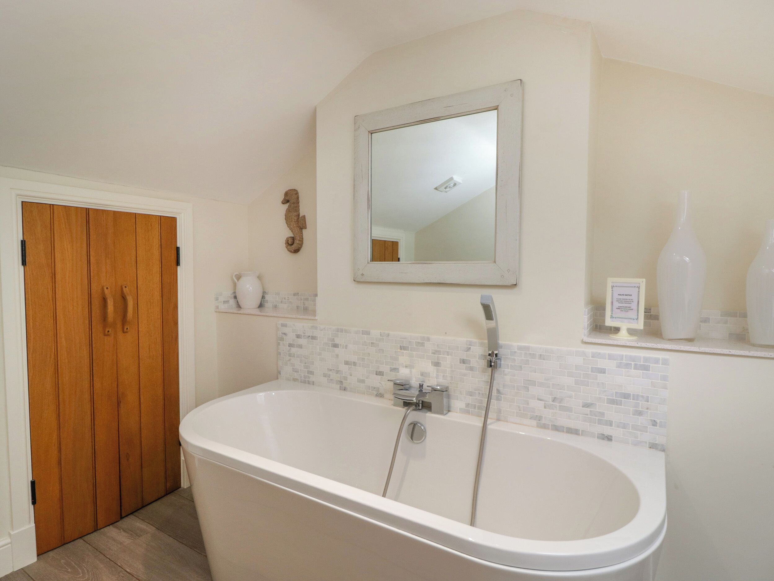 Un baño con una bañera independiente, una puerta de madera, un espejo y un estante pequeño con objetos decorativos.