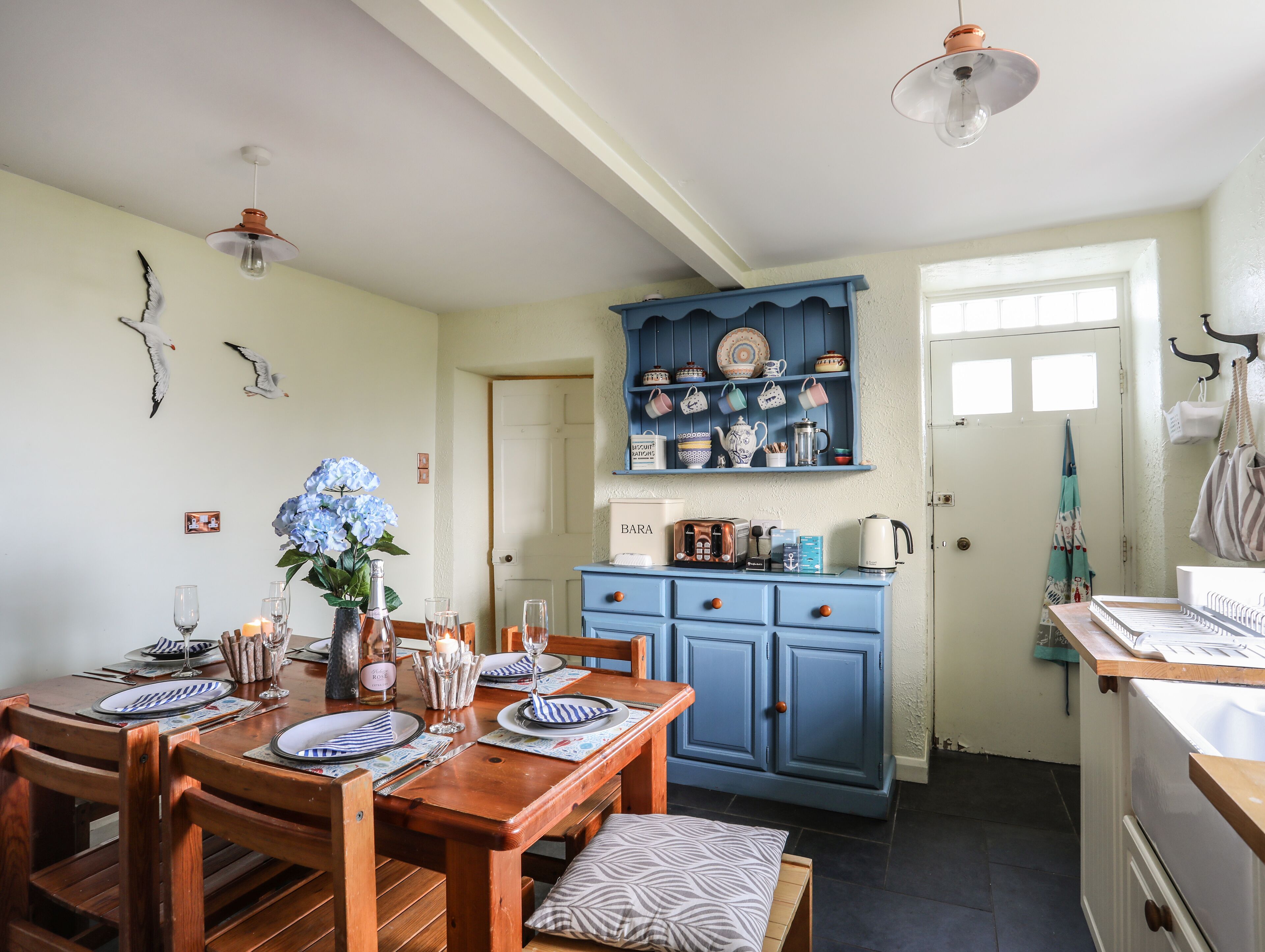 Un comedor con una mesa de madera dispuesta para cuatro personas, un armario azul con vajilla, y una zona de cocina con un refrigerador blanco.