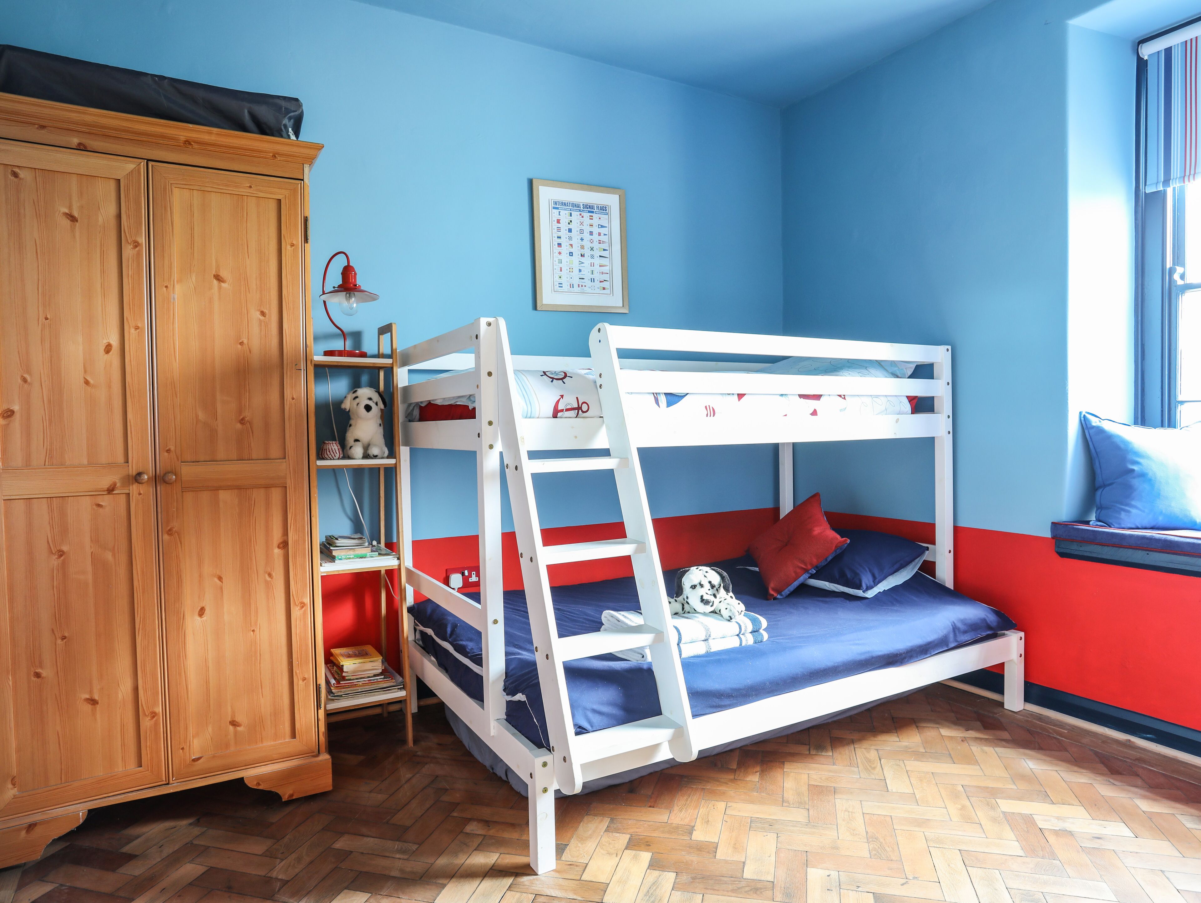 A bunk bed with a red and blue color scheme, wooden wardrobe, and a window with a view.