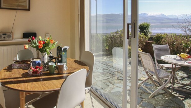 Un coin repas avec une table dressée pour deux personnes, un vase de fleurs et une vue sur l’océan par une porte vitrée.