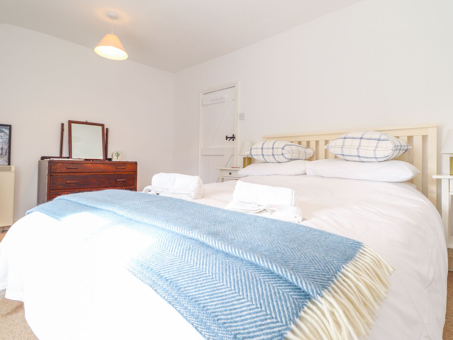 Un lit bien fait, recouvert d’une couverture bleue et blanche, une commode en bois, un miroir et une table de chevet avec une lampe.