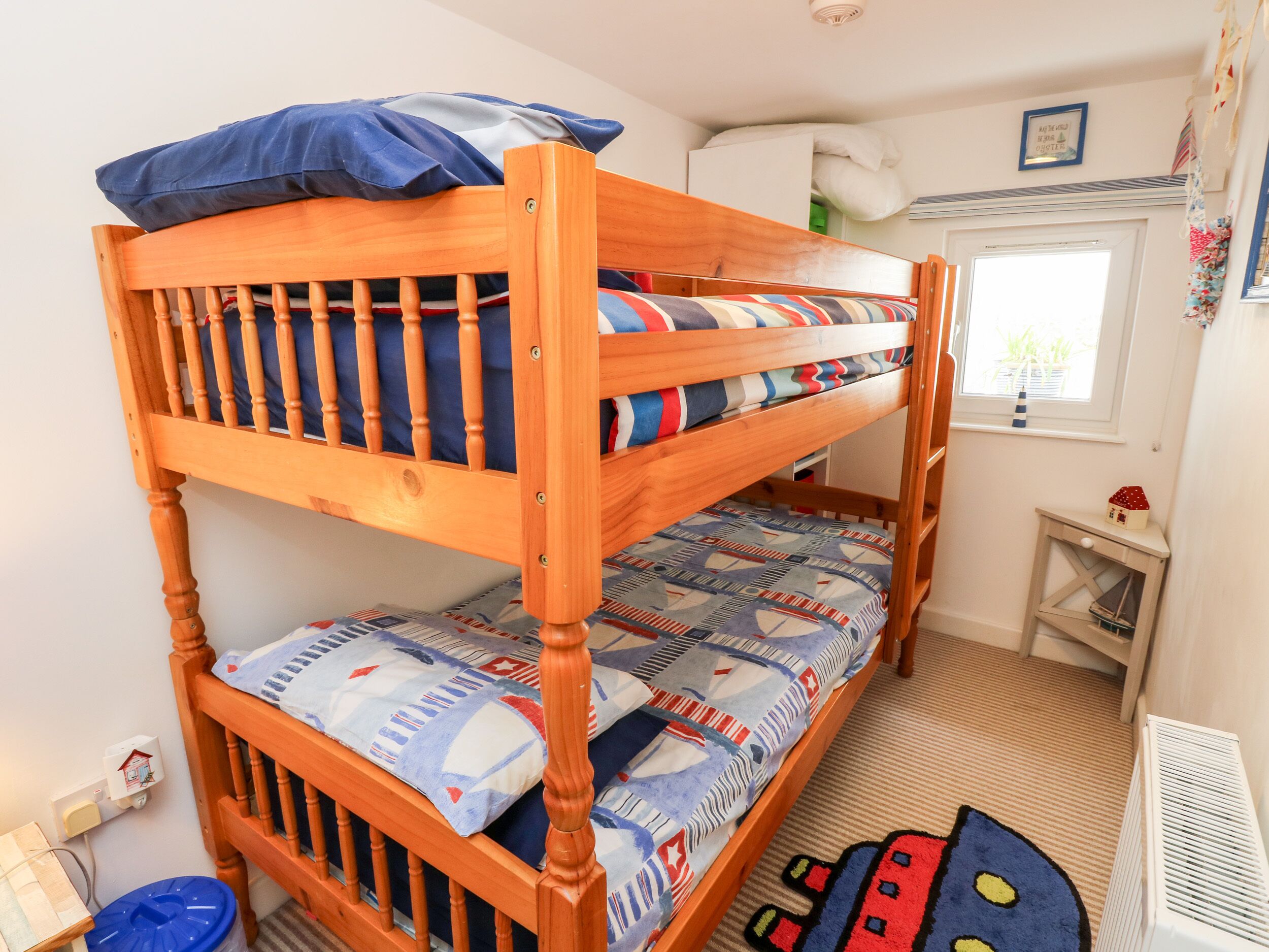 Una cama individual con ropa de cama azul y blanca, una mesita de noche con un objeto con forma de casa y una ventana con vistas a zonas verdes.