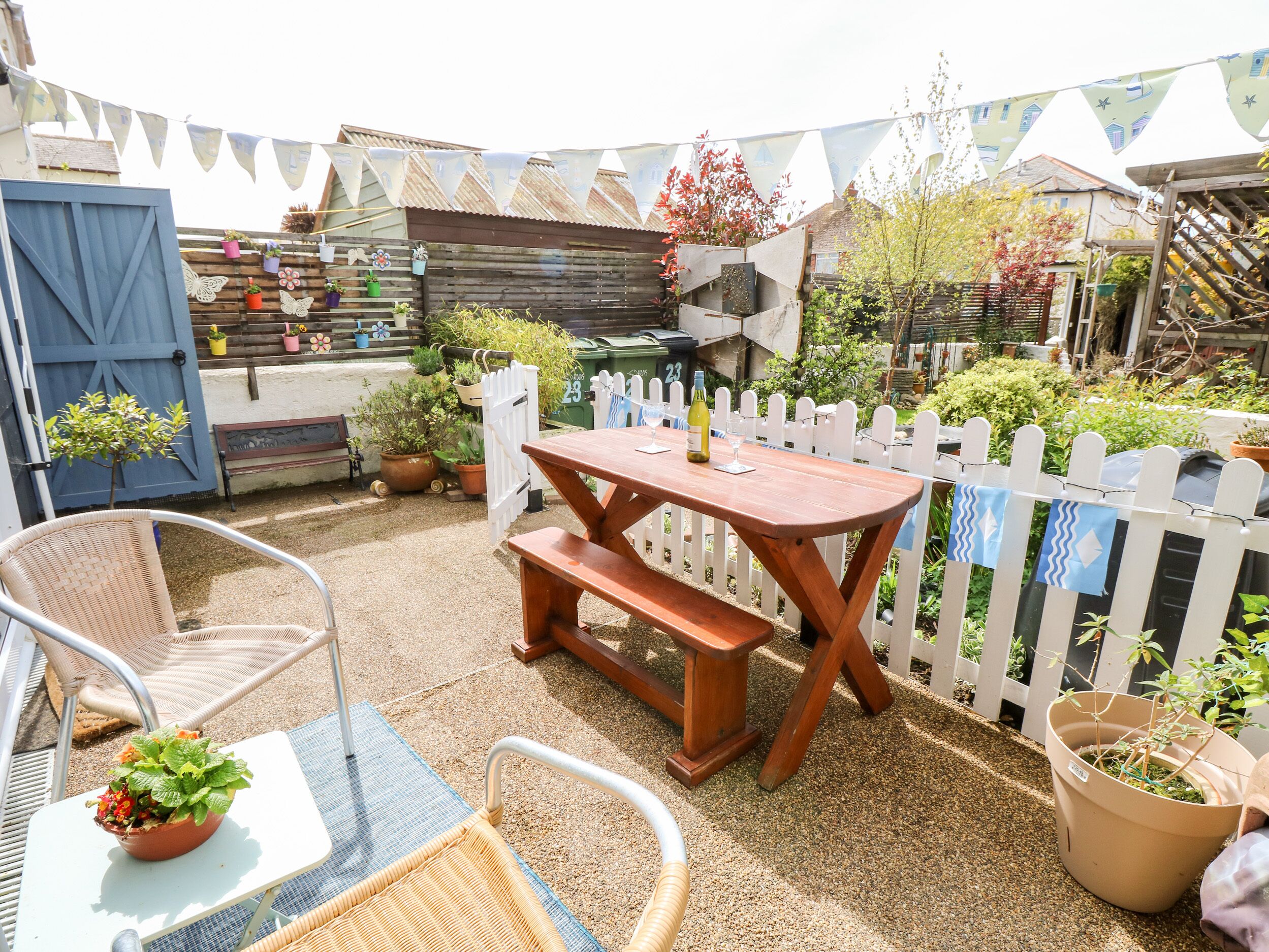 A patio area with a wooden table and chairs, surrounded by potted plants and a white picket fence.