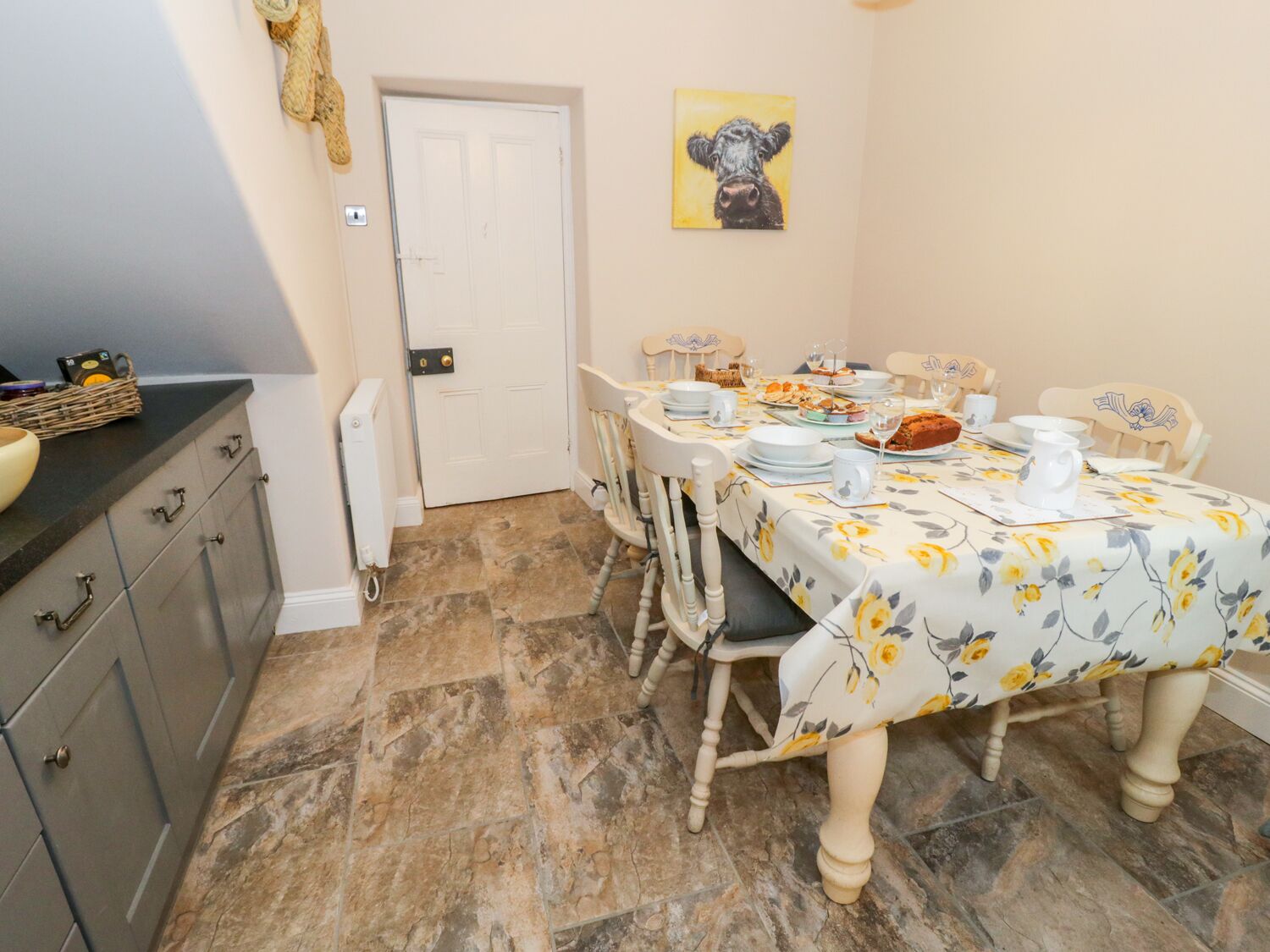 Een eetgedeelte met een tafel voor vier personen, met een tafelkleed met bloemen en witte stoelen. Op de muur hangt een schilderij van een koe, en een mand met spullen staat op het aanrecht.
