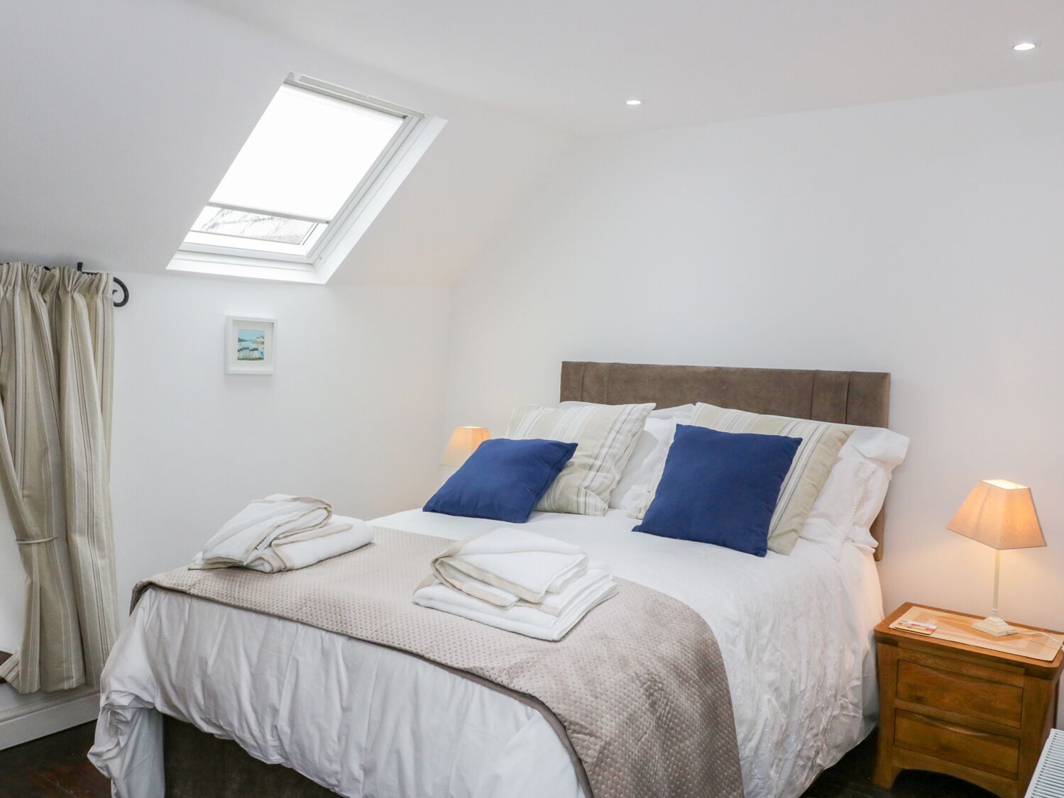 Ein ordentlich bezogenes Bett mit weißen und blauen Kissen, ein Nachttisch aus Holz mit einer Lampe und ein Oberlicht über dem Bett.