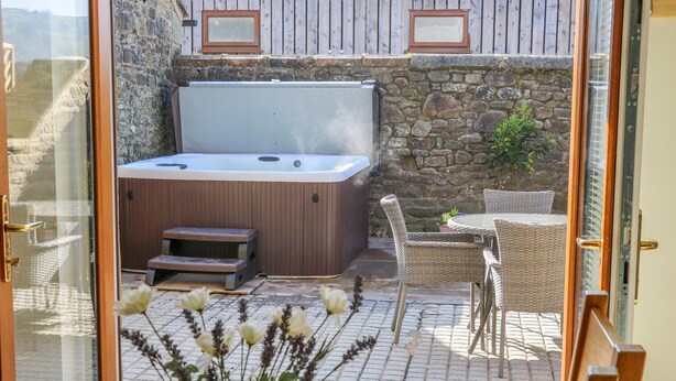 A hot tub with a wooden bench, wicker chairs, and a table with a vase of flowers in a glass-enclosed patio. - Stable View Cottage (Clitheroe)