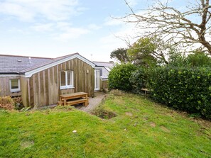 Cozy cottage with a gabled roof and attached shed, featuring a single window and door, set amidst a lush lawn and hedge.