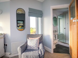 A room with a grey armchair, a window with blue blinds, a radiator, and a bathroom with a glass shower and a white sink. - Min Y Mor Trearddur Bay (Holyhead)