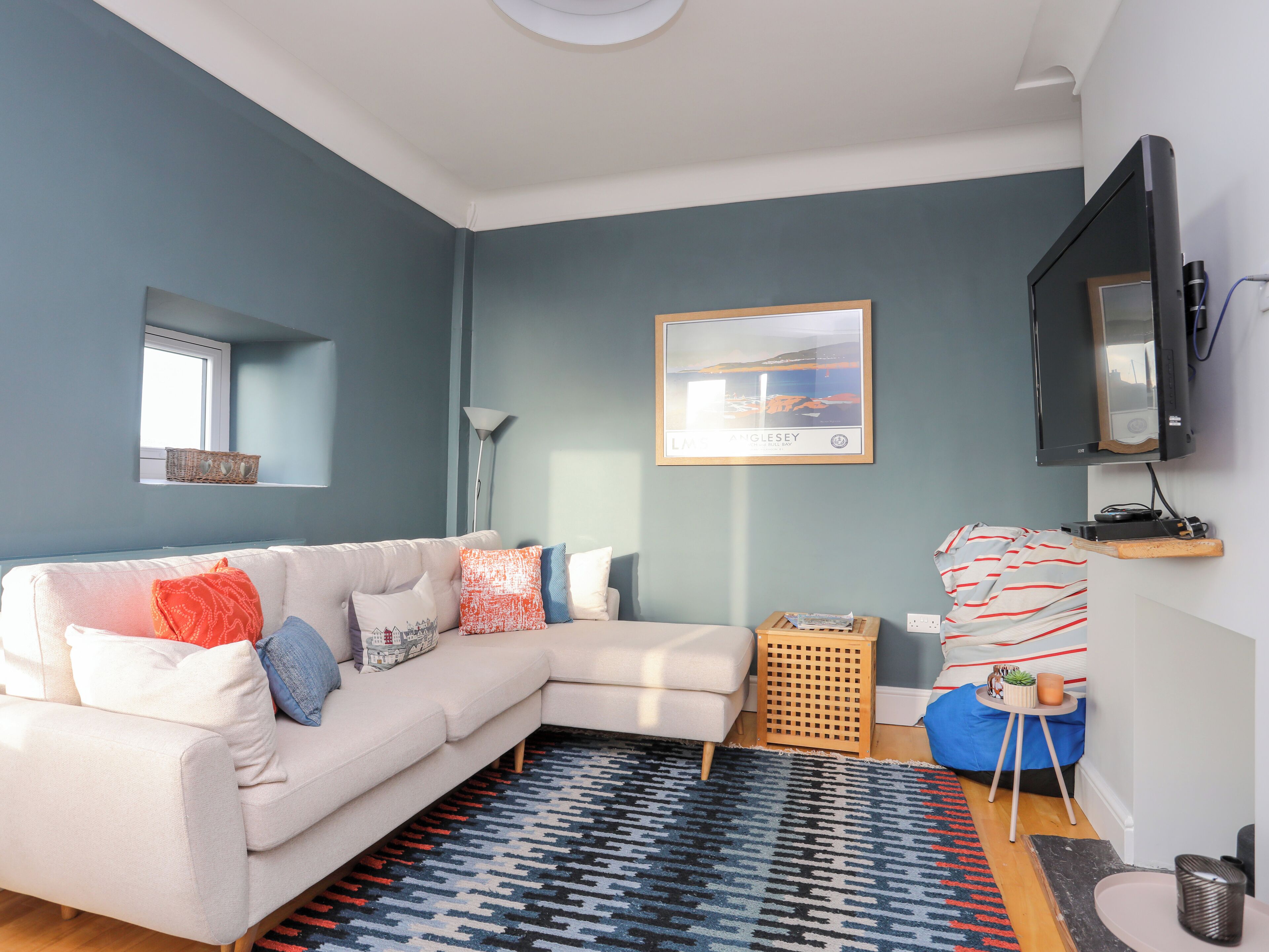 A modern living room with a light-colored sectional sofa, a patterned rug, a framed picture on the wall, and a television mounted on the wall.