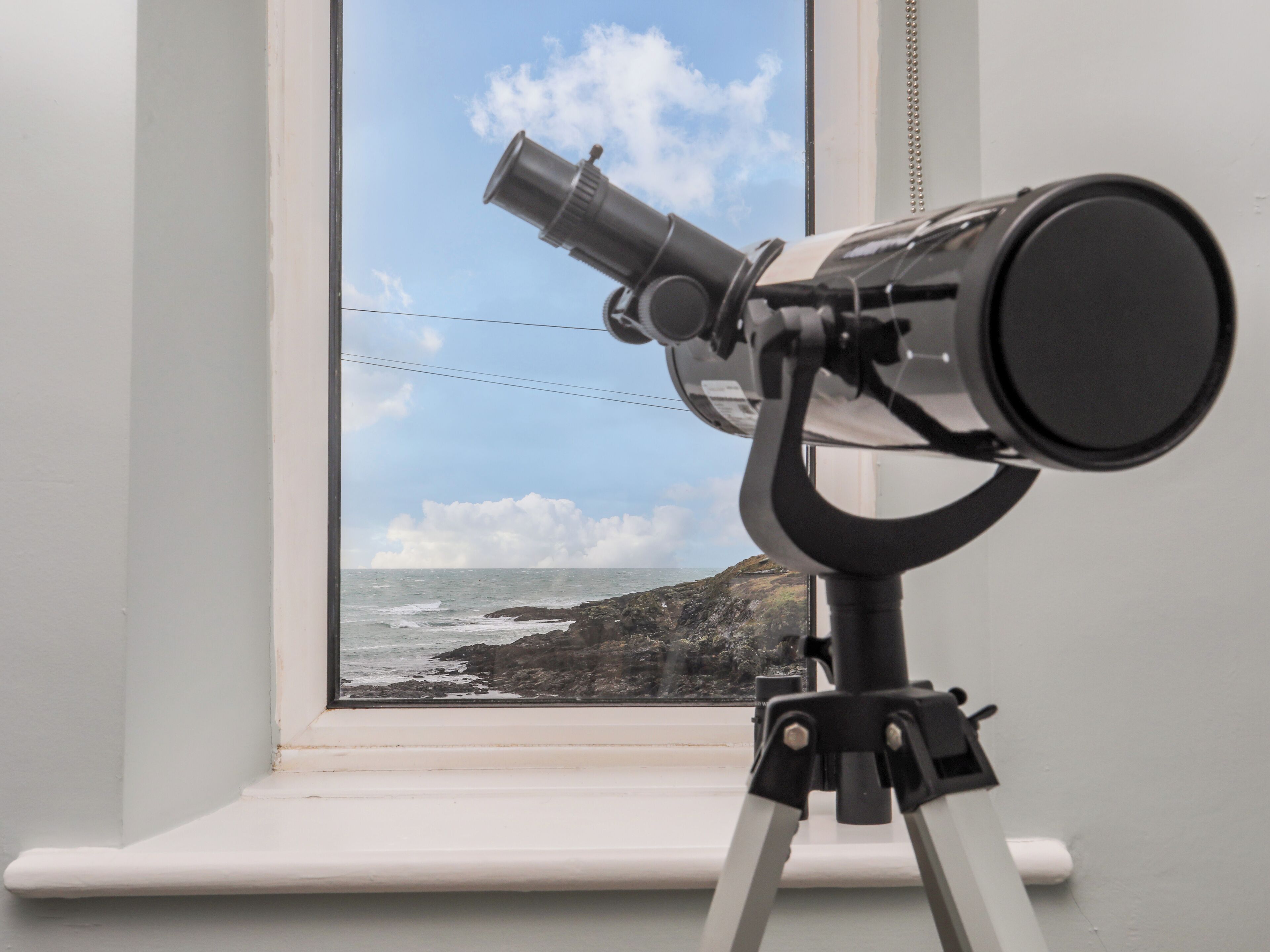 Un telescopio sobre un trípode junto a una ventana con vista a un litoral rocoso.