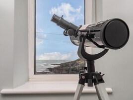 Un telescopio sobre un trípode junto a una ventana con vista a un litoral rocoso.