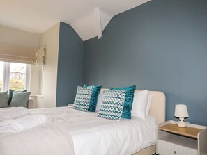 A bedroom with a bed, two patterned pillows, a nightstand with a lamp, and a window with a view of buildings. - Min Y Mor Trearddur Bay (Holyhead)