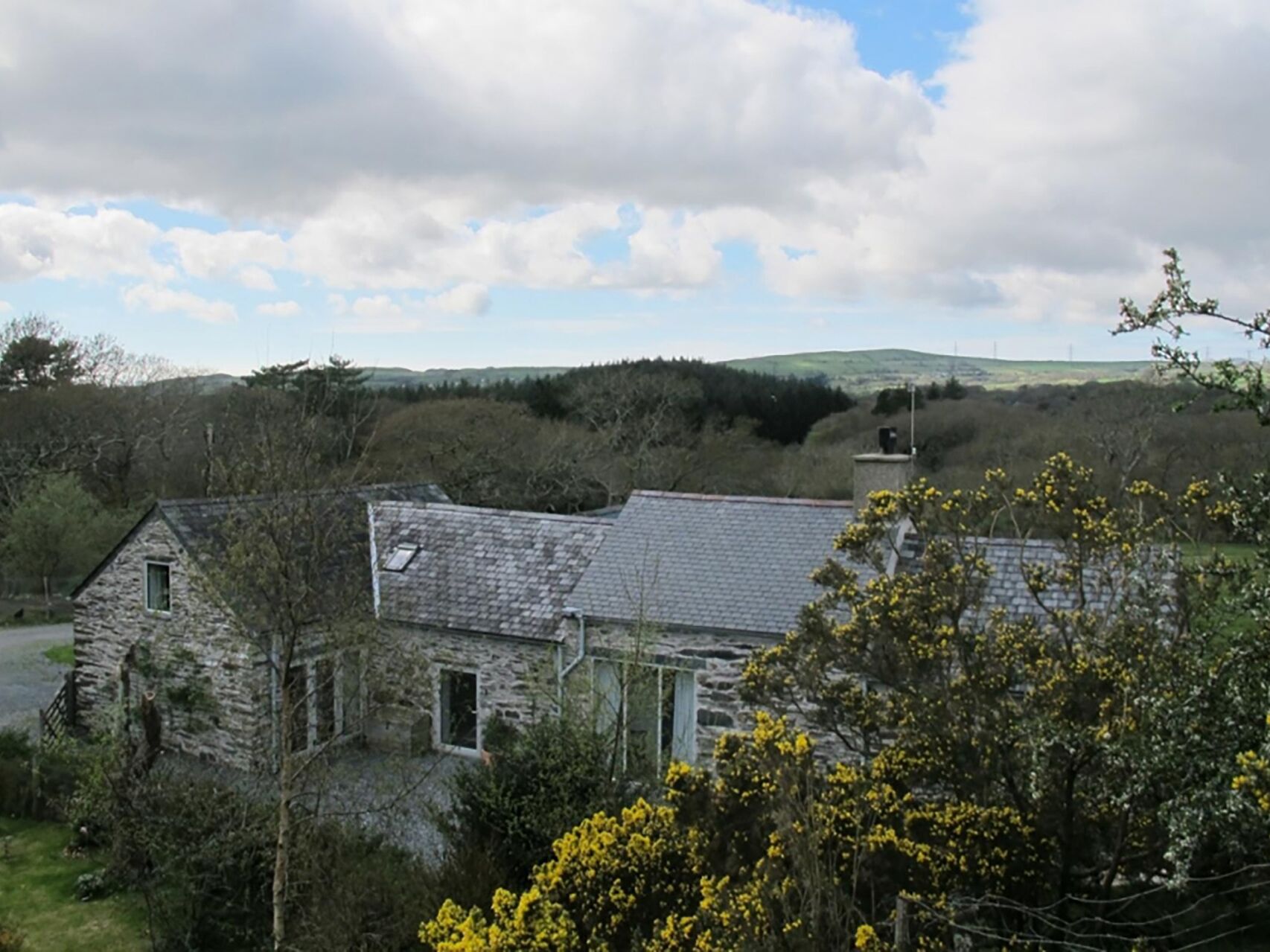 A stone building with a sloped roof, surrounded by trees and a hilly landscape.
