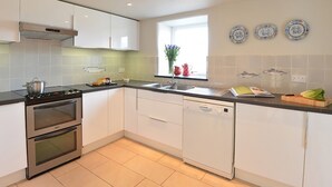 A modern kitchen with white cabinets, a stove, oven, dishwasher, and a window with decorative plates. - Cefn Isaf (Porthmadog)