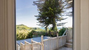 Une vue depuis une fenêtre donnant sur un escalier en bois menant à un balcon au sol en pierre, entouré de verdure et surplombant un ciel dégagé.